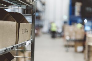 how to become an Amazon seller: image depicts a a cardboard box in focus on a shelf, with a distribution centre full of people working blurred in the background