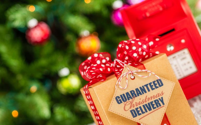 Optimise your Christmas logistics: Image shows a box tied up with a red ribbon in the foreground, with a label attached to it saying "Guaranteed Christmas Delivery". Out of focus in the background is a Christmas tree and a red post box.