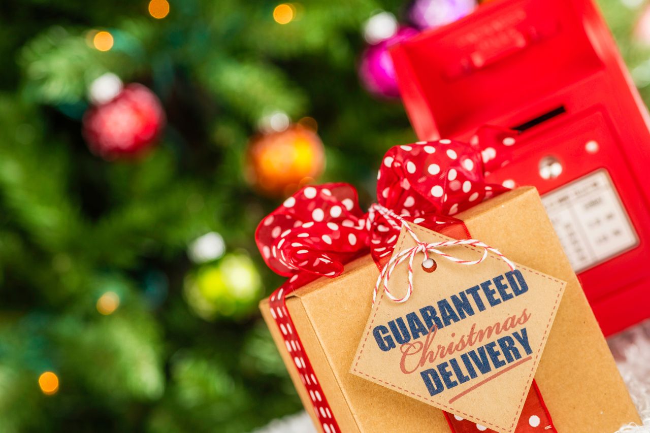 Optimise your Christmas logistics: Image shows a box tied up with a red ribbon in the foreground, with a label attached to it saying "Guaranteed Christmas Delivery". Out of focus in the background is a Christmas tree and a red post box.